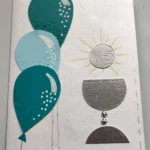 Biglietto di auguri fatto a mano per la Prima Comunione con tre palloncini nelle sfumature del blu e del turchese a sinistra, e i simboli dell'eucaristia (calice d'argento e ostia con scritta IHS) a destra su sfondo bianco materico.